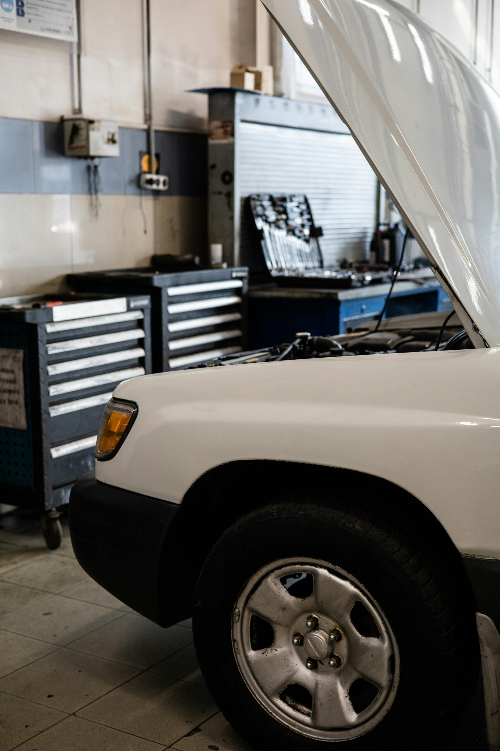 Home White car with hood open in a professional auto repair shop. Toolbox and equipment visible.